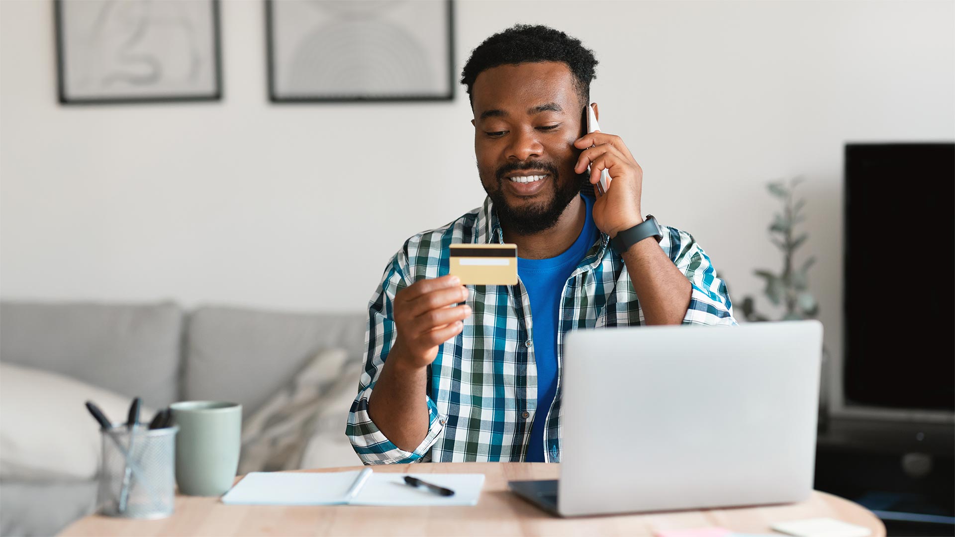 Cheerful Black Guy Holding Credit Card Talking On Phone Indoors
