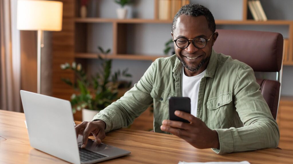 Glad busy handsome adult black businessman in glasses with computer typing on smartphone in home office interior.