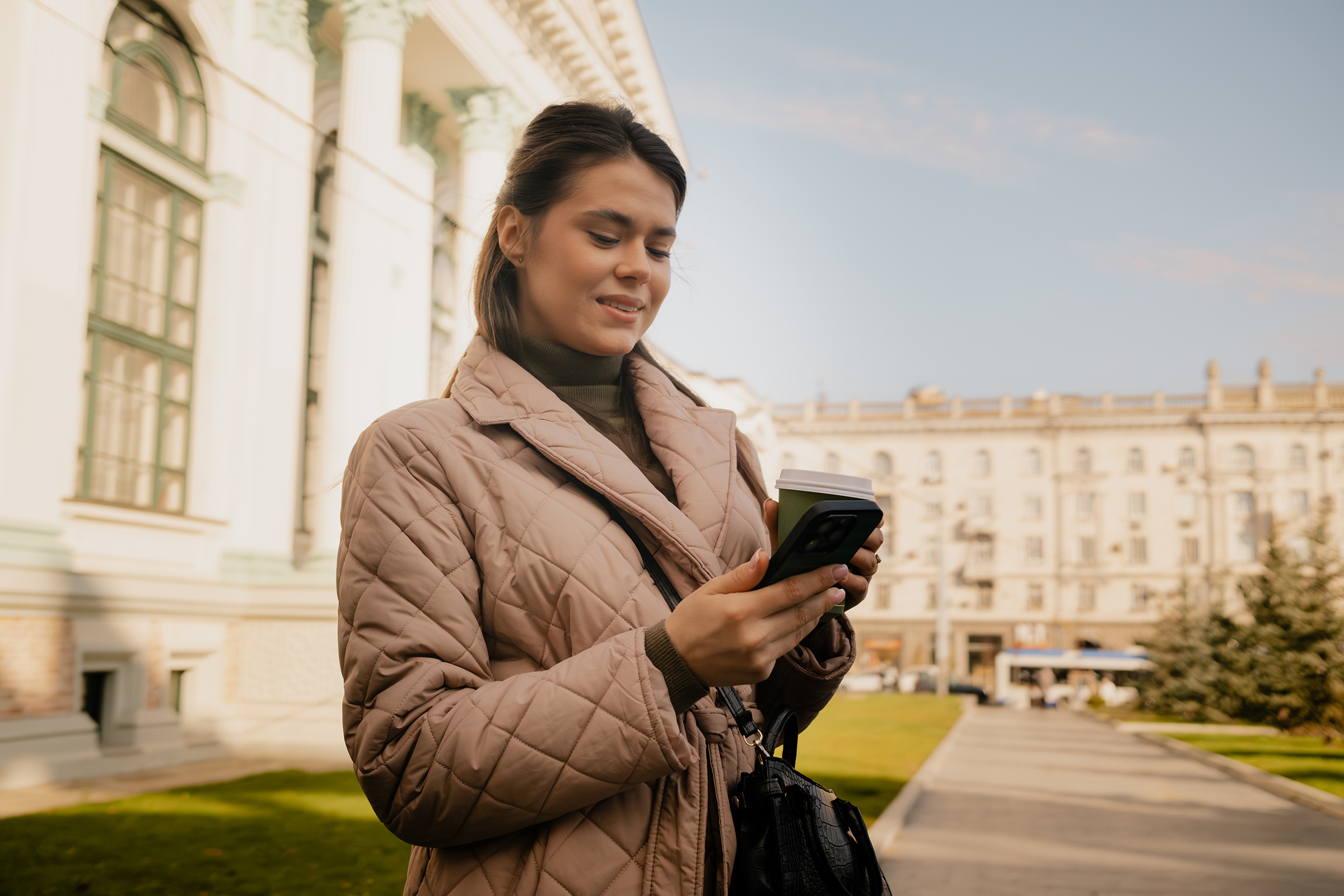 Woman checks her phone outdoors in a beige quilted jacket near a historic building